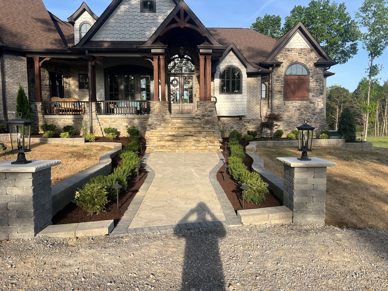Stone walkway entrance with paver pillars, landscape lighting, and planting beds leading to a stone home entry by Jordan's Landscaping